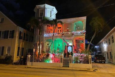Festively lit two-story Victorian house at night with wraparound porch and balcony glowing red and green, string lights along the railing, an American flag, palm trees at the entrance, and a white fence in a tropical coastal neighborhood.