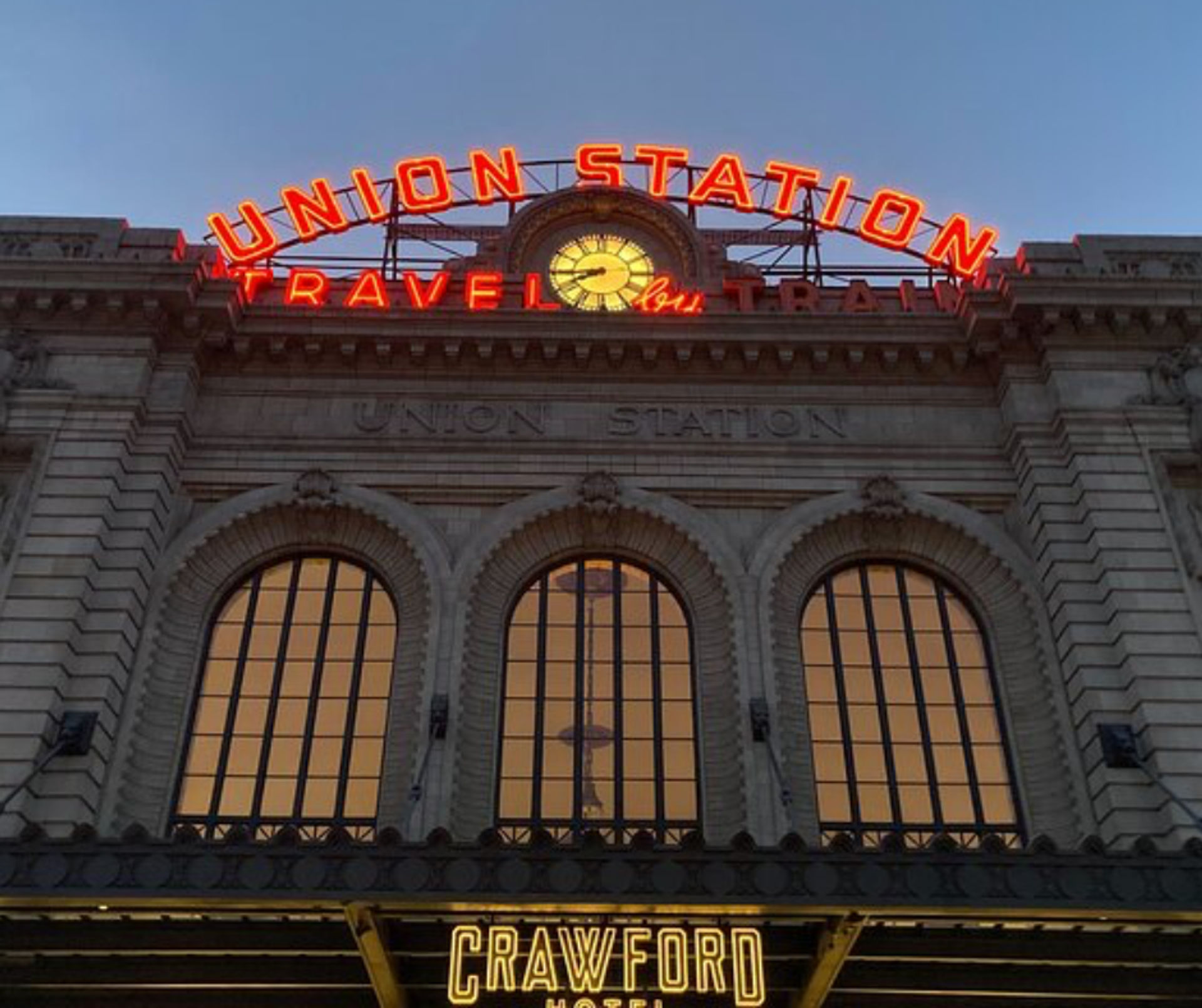 Historic downtown train station façade at dusk with glowing red neon sign and illuminated clock above three tall arched windows and a lit hotel marquee below