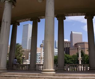 Neoclassical stone columns framing downtown skyscrapers and a tree-lined park under a clear blue sky, with a lone person seated on the balustrade.