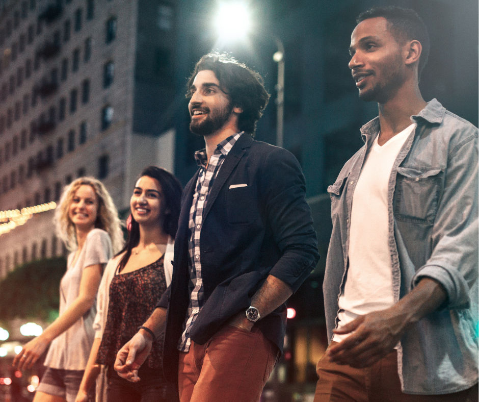 Four friends strolling together on a downtown city street at night, smiling under urban lights with tall buildings in the background.