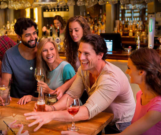 Cheerful group of five friends laughing and sharing wine, beer and cocktails at a lively neighborhood bar with warm lighting and a wooden countertop.