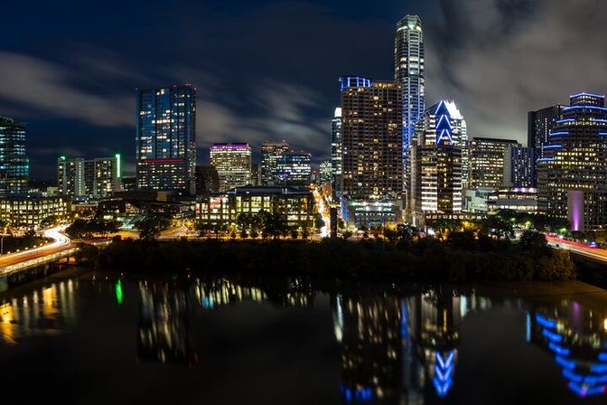 Downtown riverfront skyline at night with illuminated skyscrapers, colorful building lights and light trails reflected in the calm water.