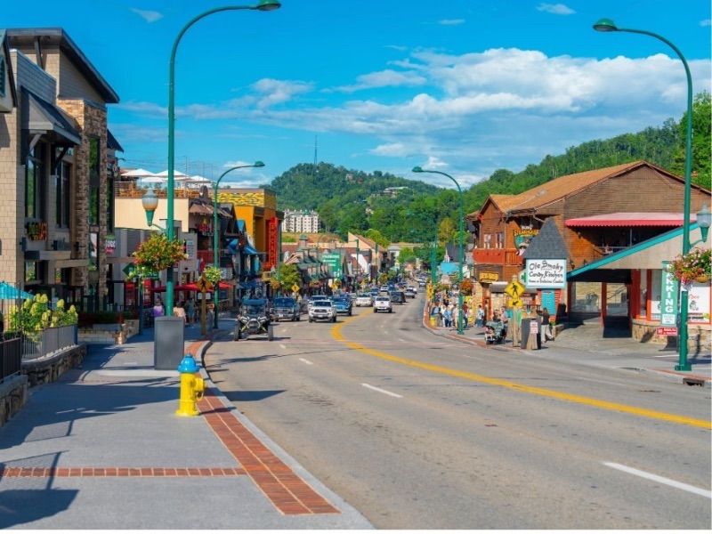 Sunny mountain-town main street lined with colorful shops and wooden storefronts, cars and strolling tourists, lamp posts and green hills in the background.