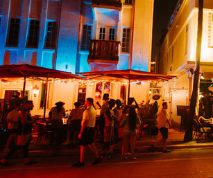 Vibrant Miami Beach nightlife scene with a crowded outdoor patio under red umbrellas, string lights and blue-orange art deco facade, people socializing and strolling on the sidewalk at night