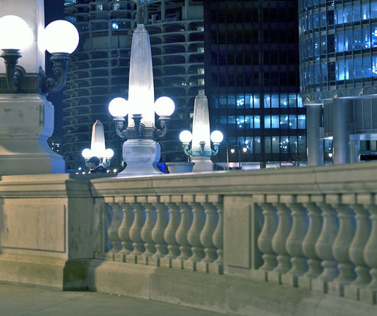 Nighttime urban bridge scene with stone balustrade, ornate glowing globe street lamps and blue-lit high-rise windows, a small pedestrian silhouette in the background.