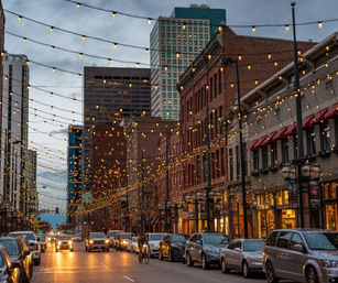 Downtown city street at dusk with twinkling string lights overhead, brick storefronts, parked cars, traffic and a bicyclist rolling through