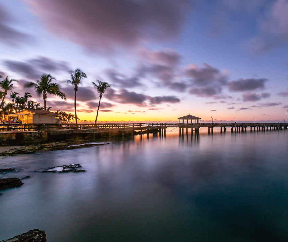 Tranquil tropical sunset over a calm coastal pier with silhouetted palm trees and a wooden gazebo, colorful clouds reflected in smooth water.