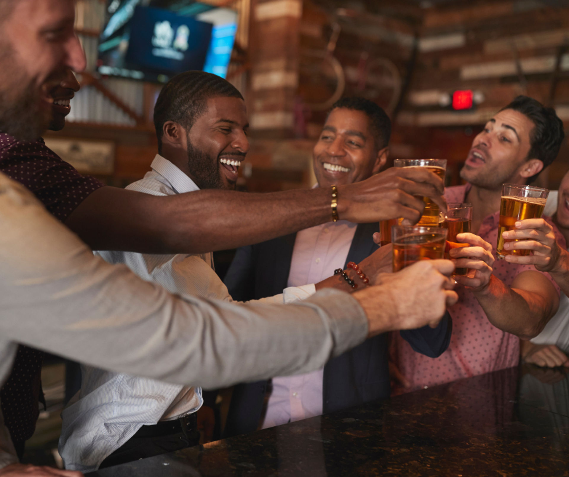 Group of friends clinking beer glasses at a rustic bar counter, laughing and celebrating under warm wood-paneled lighting.