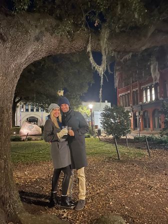 Smiling couple in winter coats and beanies holding drinks under a moss-draped oak tree in a lit historic downtown square at night, with brick buildings in the background.