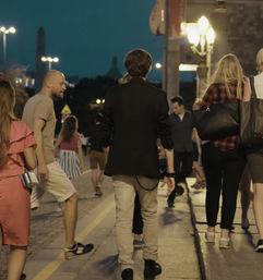 Evening urban street scene with pedestrians strolling along a lit sidewalk under glowing lampposts, a young man in a black jacket walking center-back, casual summer outfits and lively city nightlife atmosphere.