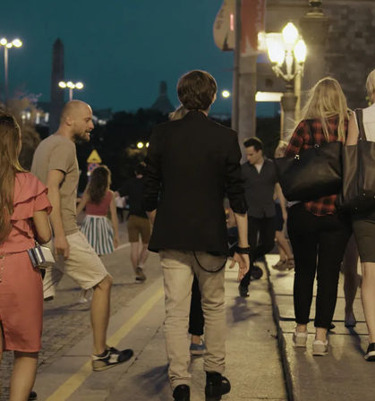 Evening urban street scene with pedestrians strolling along a lit sidewalk under glowing lampposts, a young man in a black jacket walking center-back, casual summer outfits and lively city nightlife atmosphere.