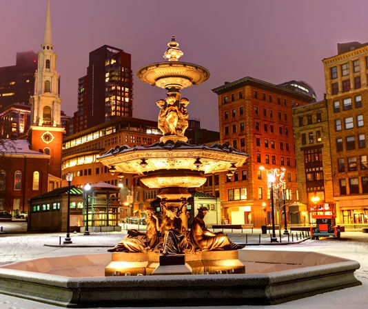 Ornate illuminated fountain in a snowy urban plaza at dusk, framed by historic brick buildings and a church steeple with warm city lights against a purple sky.