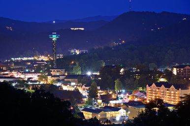 Twilight view of a mountain resort town lit up at night, featuring a spiral observation tower, hotels and shops clustered in a valley with forested hills in the background.