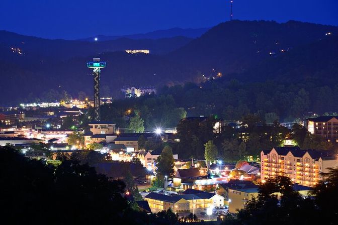 Twilight view of a mountain resort town at night, sparkling hotel lights and shops clustered in a valley with a glowing observation tower rising above forested hills.