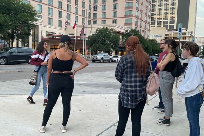 Group of casually dressed young adults standing and chatting on a downtown sidewalk in front of a multi-story hotel with flags and parked cars.