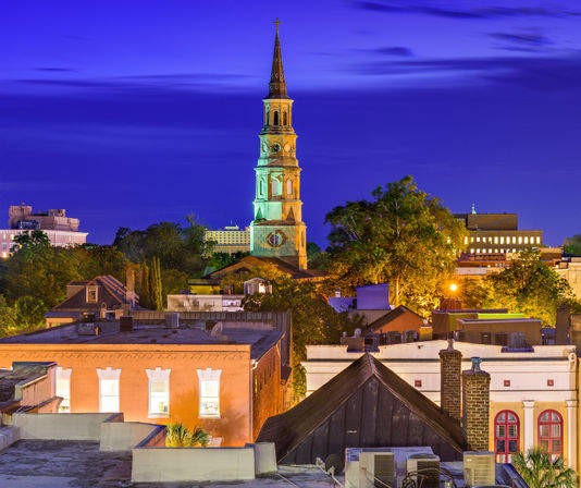 Twilight cityscape with an illuminated historic church steeple rising above rooftops, leafy trees, and warm streetlights under a vibrant indigo sky.