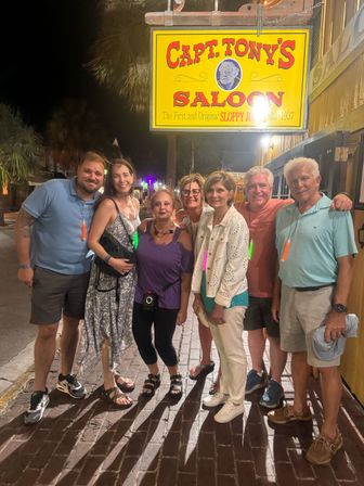 Seven friends posing on a brick sidewalk at night beneath a bright yellow vintage saloon sign, palm trees and neon-lit coastal downtown street in the background