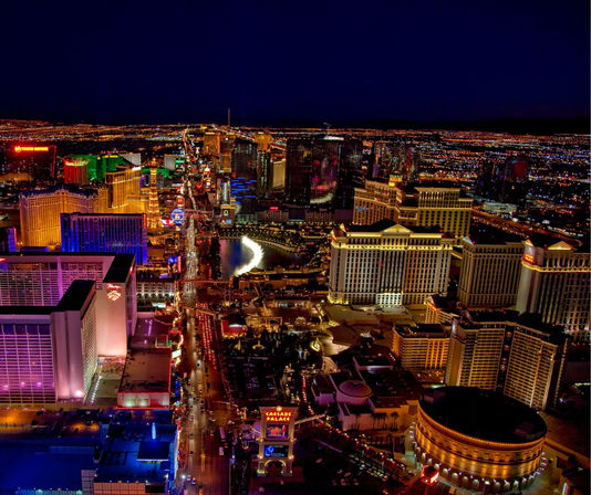 Aerial night view of the glittering Las Vegas Strip in Nevada, with brightly lit hotels and casinos, neon signs, a glowing fountain and a busy illuminated boulevard.