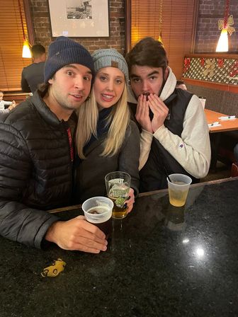 Three friends in winter beanies smiling at a cozy pub bar counter, holding beer glasses against a brick wall with holiday lights.