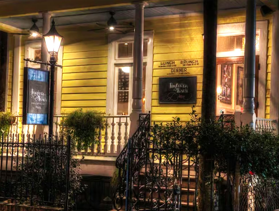 Cozy evening view of a yellow wooden restaurant porch with a vintage street lamp, wrought-iron gate, hanging menu sign and potted greenery.