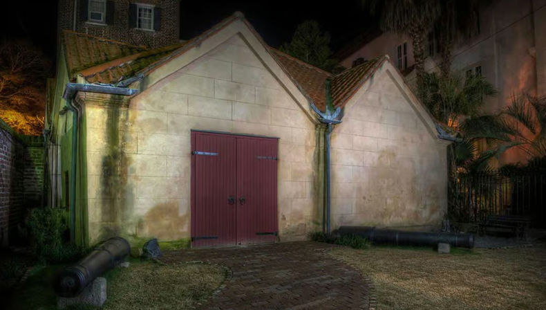 Night view of a historic stone outbuilding with red double doors and tiled gabled roofs in a moody courtyard, flanked by antique cannons, brick pathway and tropical plants.
