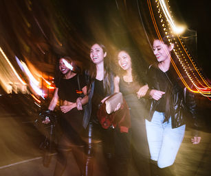 Four friends laughing arm-in-arm on a downtown city street at night, motion-blurred streaks of light and leather jackets capturing a lively urban nightlife scene.