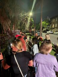 Nighttime walking tour group gathered by an ornate iron fence beneath Spanish moss on a tree-lined city sidewalk, lit by streetlights with parked cars along the road.