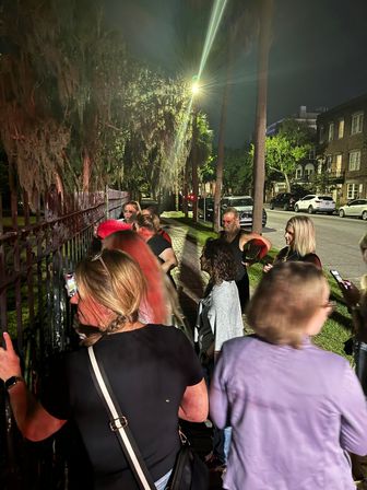 Nighttime walking tour group gathered by an ornate iron fence beneath Spanish moss on a tree-lined city sidewalk, lit by streetlights with parked cars along the road.