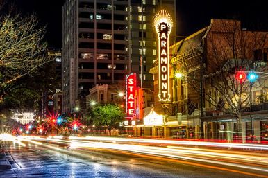 Nighttime downtown street scene with historic theater marquees, wet pavement reflections and colorful long-exposure car light trails.
