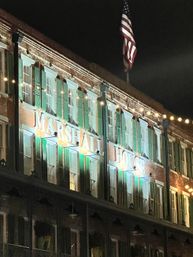 Illuminated historic brick hotel facade at night with green shutters, warm string lights, and an American flag overhead.