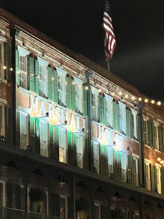 Illuminated historic brick hotel facade at night with green shutters, warm string lights, and an American flag overhead.