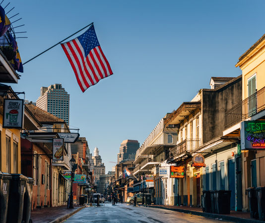 Sunlit New Orleans French Quarter street with an American flag overhead, colorful balconies, neon bar signs, and downtown skyline in the distance