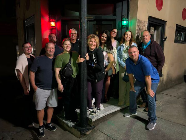 Eleven adults smiling in a nighttime urban group photo on a sidewalk outside a doorway lit by red and green lanterns, casual clothing and lively downtown nightlife vibe.