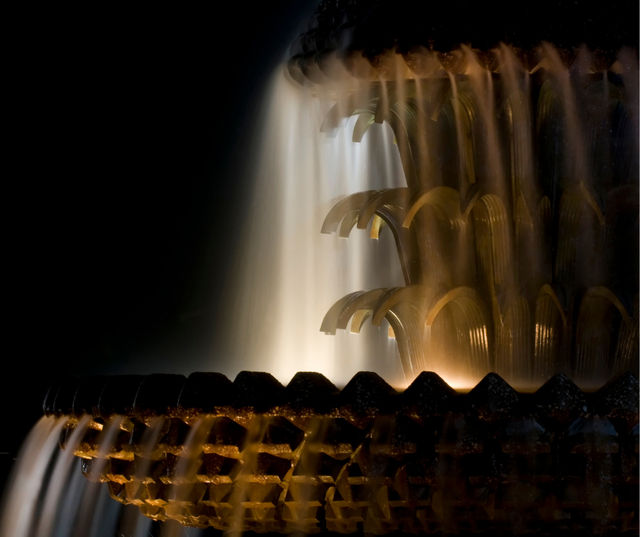 Close-up of a bronze-toned tiered fountain at night, warm lighting illuminating silky cascading water streams against a dark background