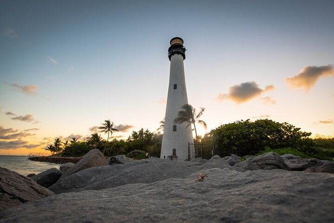 Tall white lighthouse on a tropical rocky shoreline at sunset, framed by palm trees, calm sea, and a pastel sky