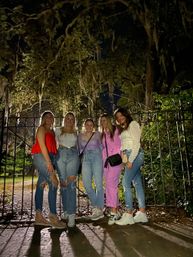 Five friends posing at night in front of an ornate iron gate beneath moss-draped trees in a park, wearing casual jeans and colorful tops, warmly lit on a paved path.