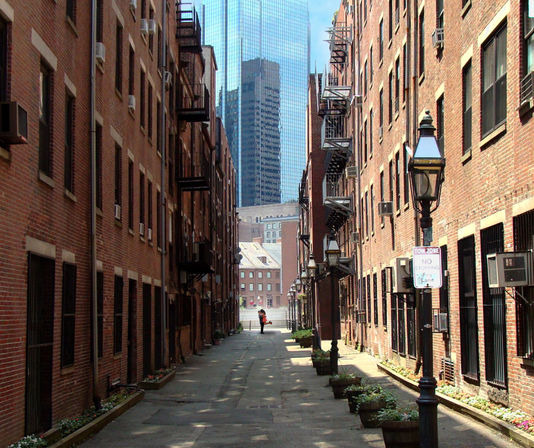 Sunlit narrow urban alley framed by red brick apartment buildings with iron fire escapes and vintage street lamps, planters along the sidewalk, a lone pedestrian at the far end and reflective glass skyscrapers beyond.