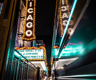 Nighttime downtown Chicago theater marquee glowing with vintage bulbs and neon, reflected on a passing bus as streaking city lights illuminate the street.