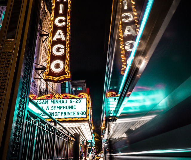 Nighttime downtown Chicago theater marquee glowing with vintage bulbs and neon, reflected on a passing bus as streaking city lights illuminate the street.