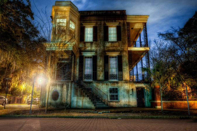 Historic two-story Southern mansion at dusk with ironwork balconies, exterior staircase and bay window, weathered stucco facade lit by warm street lamps and surrounded by trees