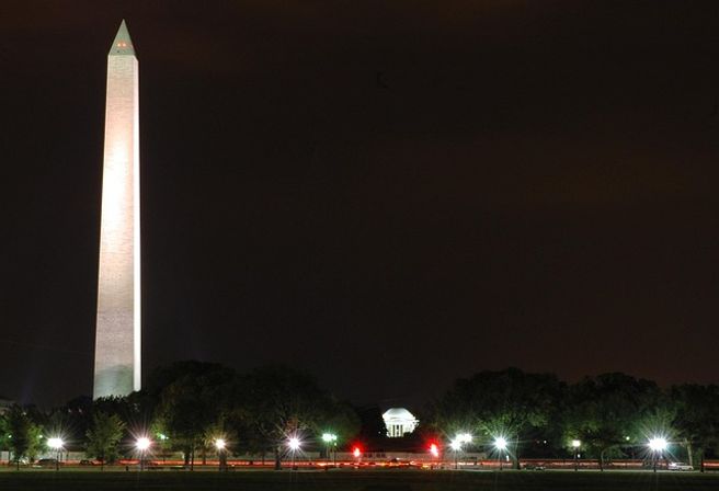 Night view of the illuminated Washington Monument with the Jefferson Memorial glowing in the distance on the National Mall in Washington, D.C., framed by trees and park lights.