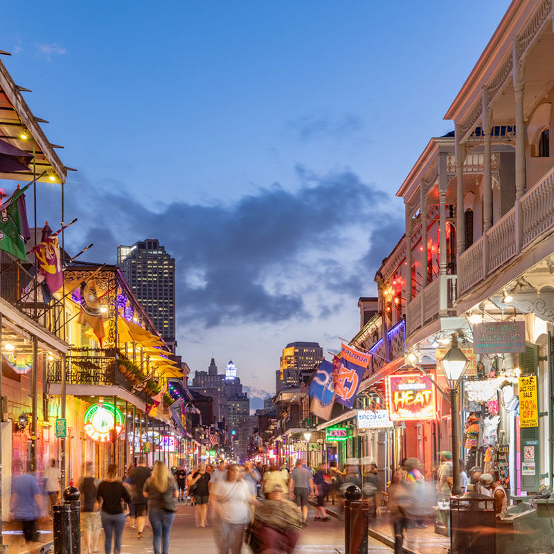 Dusk on a lively New Orleans French Quarter street, crowds strolling under wrought-iron balconies, colorful neon signs, flags and warm streetlights.