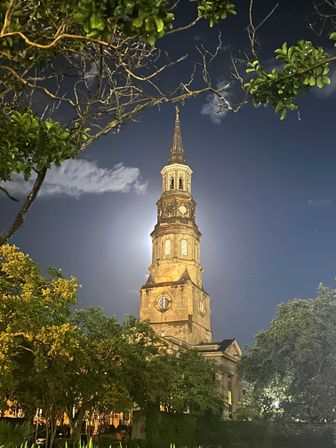 Moonlit historic church steeple and clock tower at night, framed by oak trees and bare branches against a deep blue sky