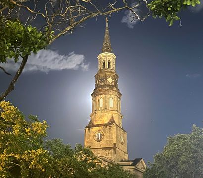 Moonlit historic church steeple and clock tower at night, framed by oak trees and bare branches against a deep blue sky