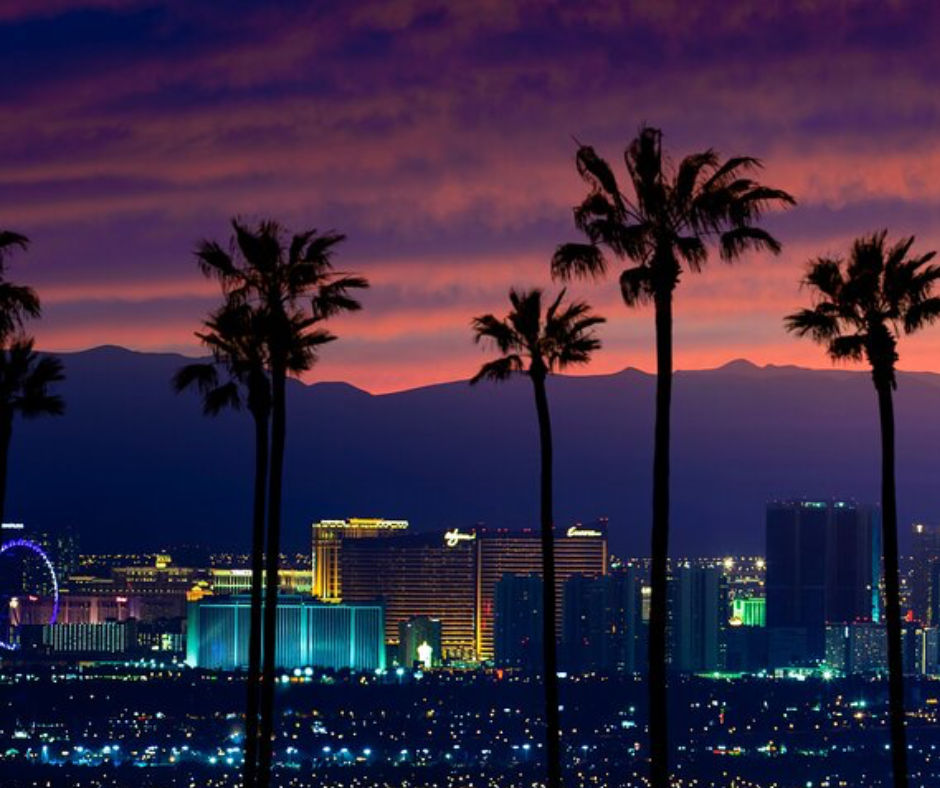Las Vegas skyline at sunset with silhouetted palm trees, colorful neon-lit hotels and a Ferris wheel framed by purple-orange mountains