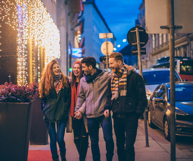 Four friends laughing and walking arm‑in‑arm on a downtown city sidewalk at dusk, wearing winter coats and scarves beneath warm holiday string lights.