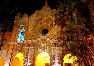 Ornate Spanish Colonial Revival façade glowing under warm night uplighting, arched entryways and intricate carvings framed by tall palm trees.