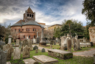 Historic cemetery with weathered tombstones and mossy grave slabs in front of a round red-brick church and bell tower, framed by oak and palm trees under a dramatic cloudy sky.