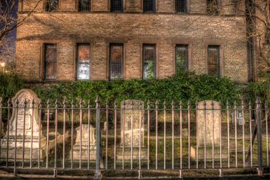 Nighttime view of a historic churchyard with weathered stone headstones behind a spiked wrought-iron fence, set against a brick building with tall leaded windows and warm evening lights.