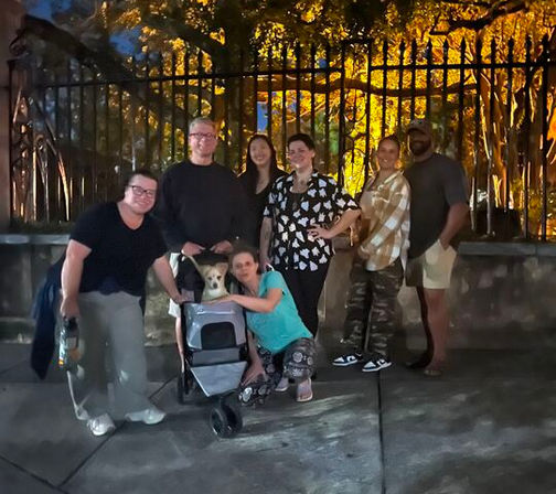Evening group photo of seven people on a city sidewalk posing with a small dog in a stroller in front of an ornate iron fence and warmly lit trees.
